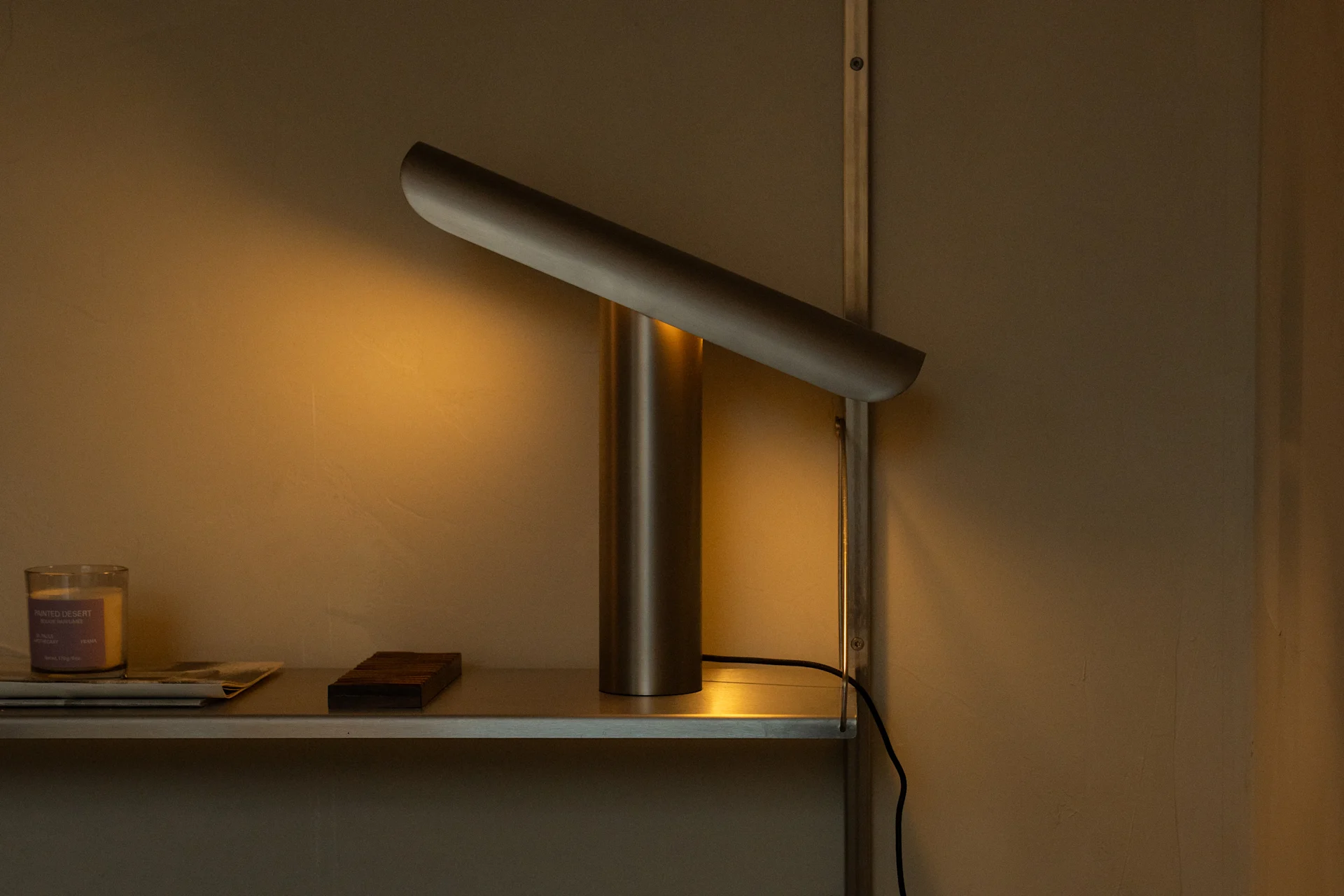 En modern metallisk lampa med cylindrisk bas och diagonal ljuslist kastar ett varmt sken på en hylla med ett ljus och papper.