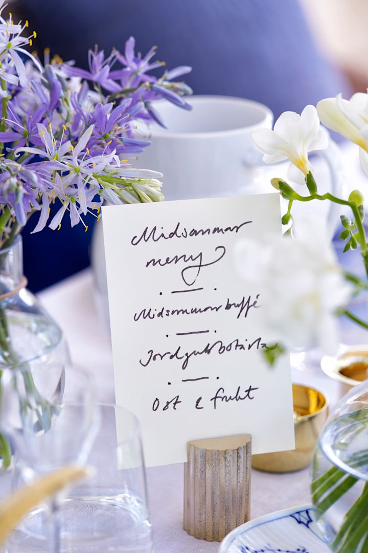 Tips till midsommardukning med handskriven midsommarmeny placerad i korthållare i mässing. 