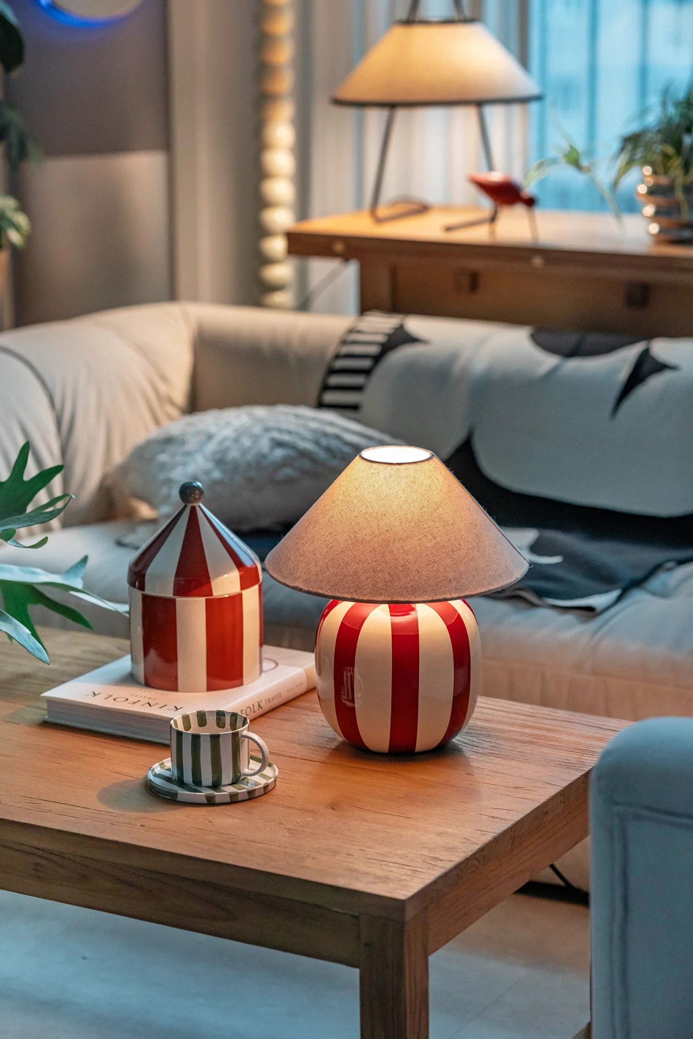 Ett mysigt vardagsrum med en röd- och vitrandig lampa, matchande burk och en grönrandig kopp på ett soffbord i trä.