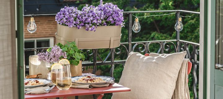Solig balkong med röda utemöbler. En beige Bau blomlåda från Ferm Living och över stolen hänger en beige pläd från ERNST. 