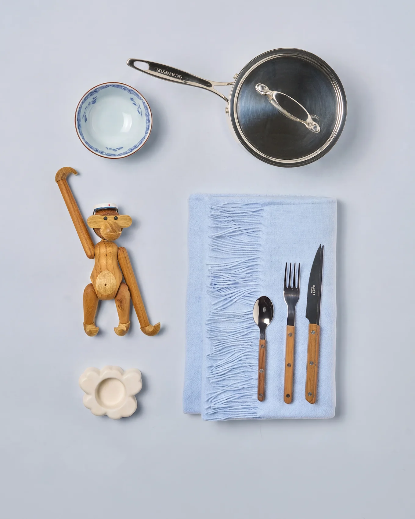 Flat lay av köksutrustning och inredning: en silverkastrull, mönstrad skål, träapa, blå filt med bestick och molnformat fat.
