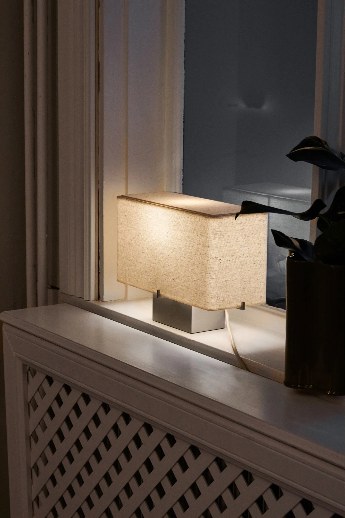 Rektangulär bordslampa med beige tygskärm och silverfot på en vit fönsterbräda, bredvid ett fönster och en växt.