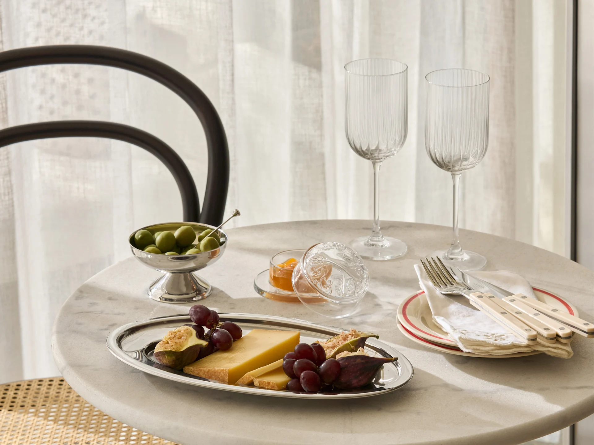 Ett marmorbord dukat med ett silverfat med ost, vindruvor, fikon, en skål oliver, sylt, två räfflade glas och bestick på tallrikar.