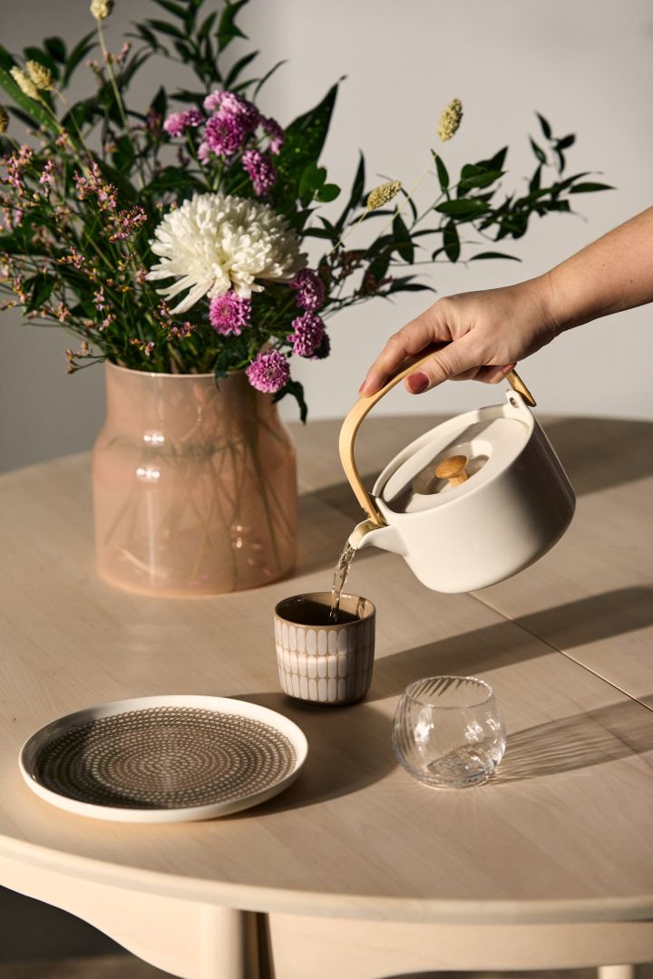 En kvinnlig arm håller i Marimekkos Oiva tekanna i vit, och häller ned kaffe från denna ner i Alku muggarna från Marimekko i färgen terra-white. 