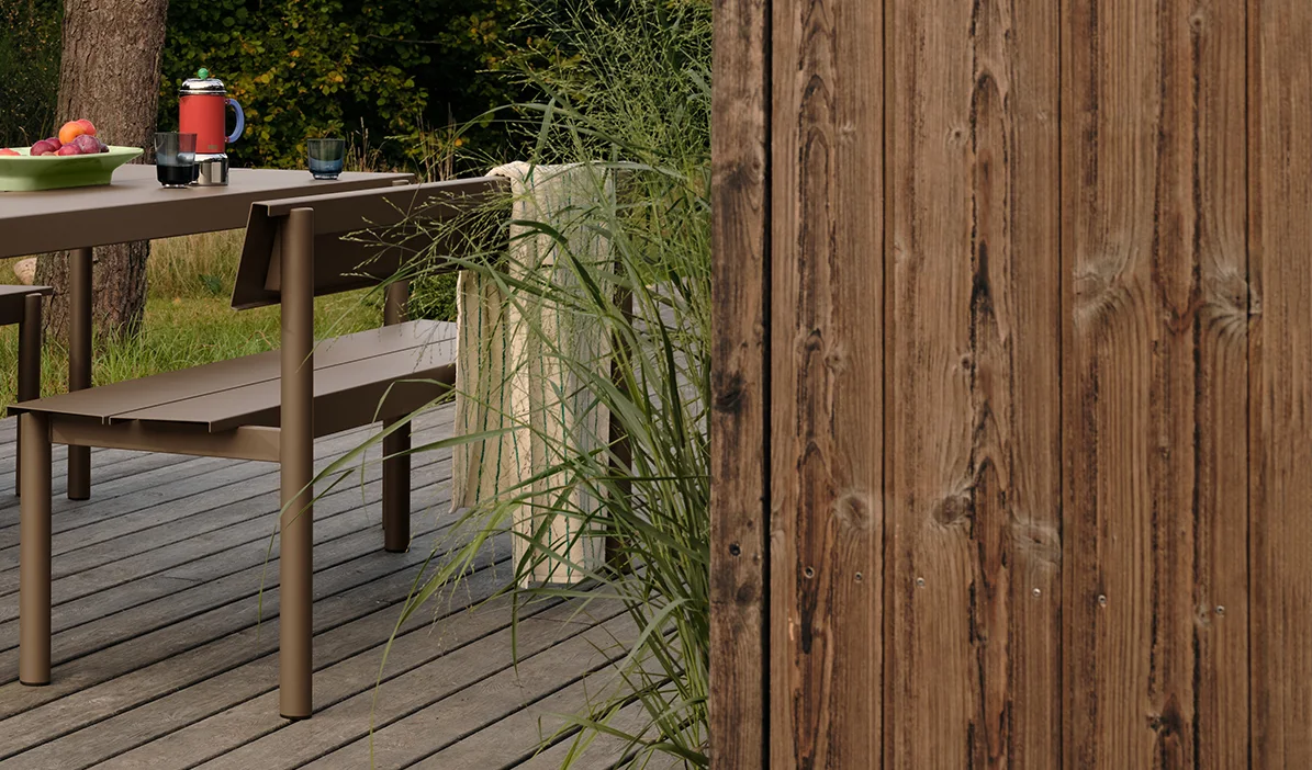 Brunt metall utomhusbord och bänk på trädäck med frukt och drycker, bredvid högt gräs och ett trästaket.