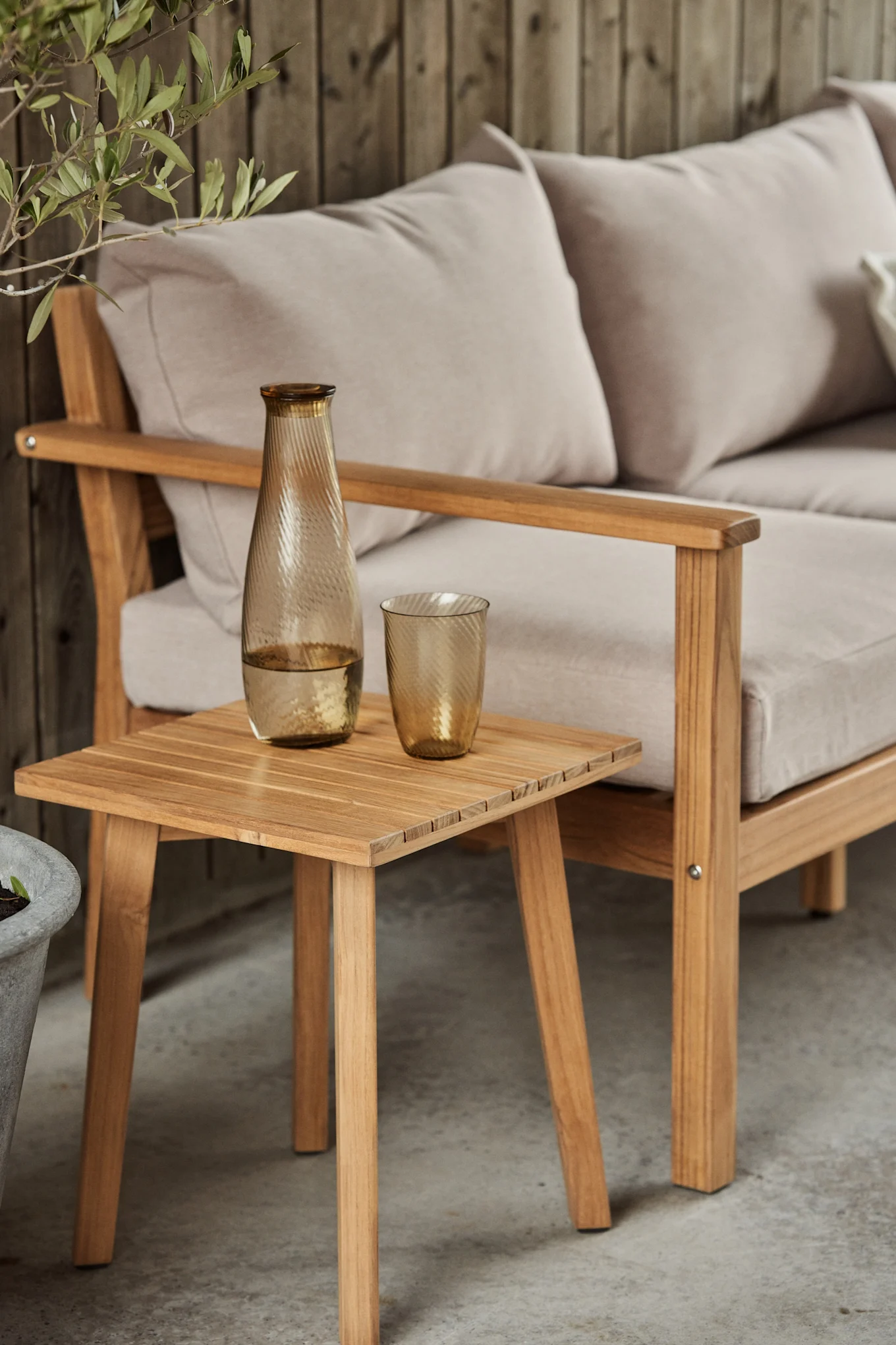 Träsoffa för utomhusbruk med ljusa dynor, ett sidobord i trä med bärnstensfärgad räfflad glaskaraff och matchande glas. Bli inspirerad att inreda din balkong 2026. 