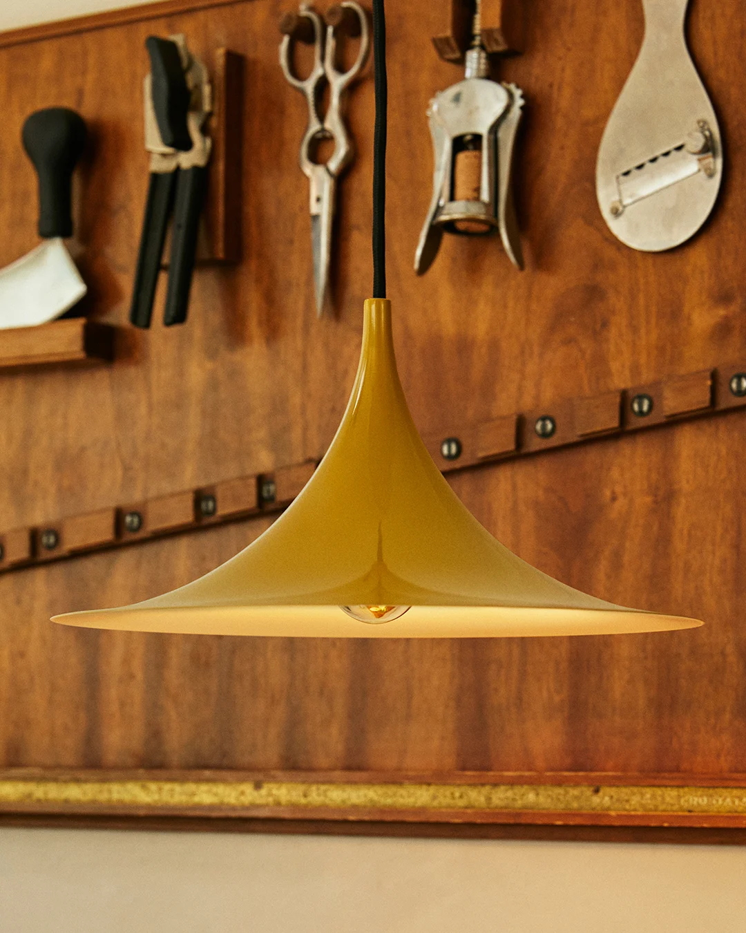 Guldfärgad klockformad taklampa med synlig glödlampa, mot en trävägg med hängande verktyg.