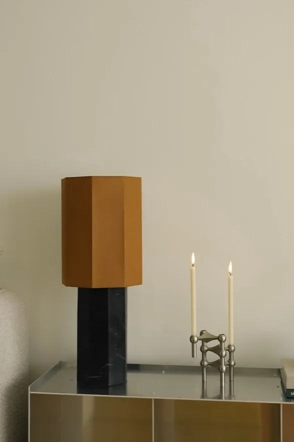 Modern bordslampa med sexkantig brun skärm och svart marmorbas, bredvid en silverfärgad modulär ljusstake med två tända vita ljus.