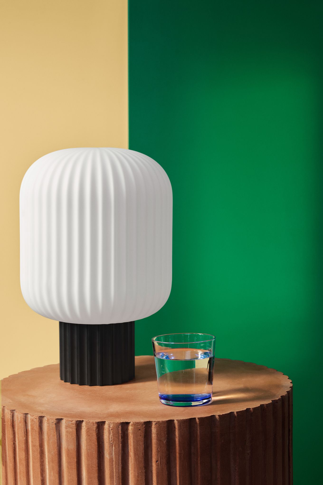 Bordslampa i räfflat glas från Broste Copenhagen mot en tvåfärgad vägg i grönt och gult. 