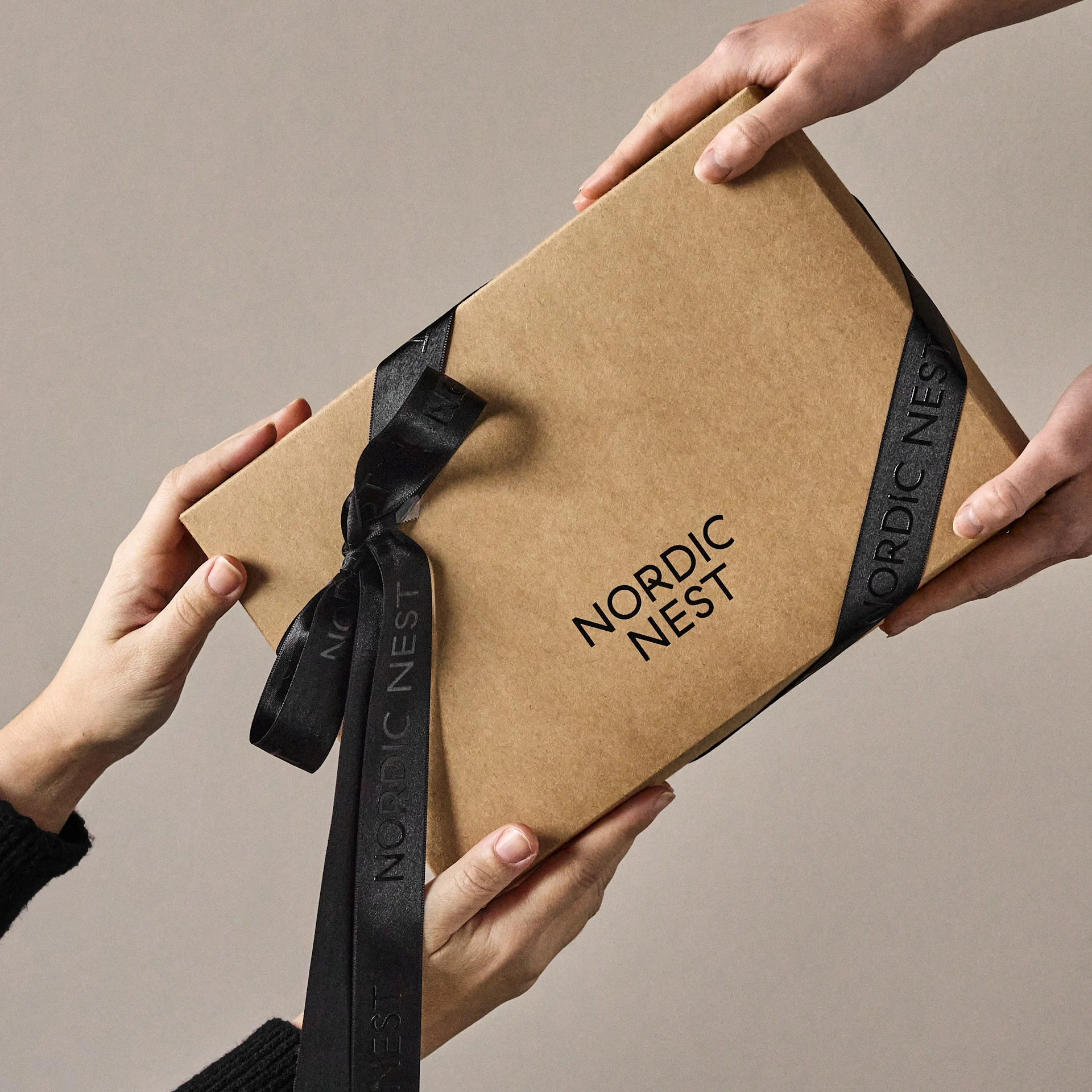 Händer som utbyter en brun presentförpackning med 'Nordic Nest'-logotyp och svart band.
