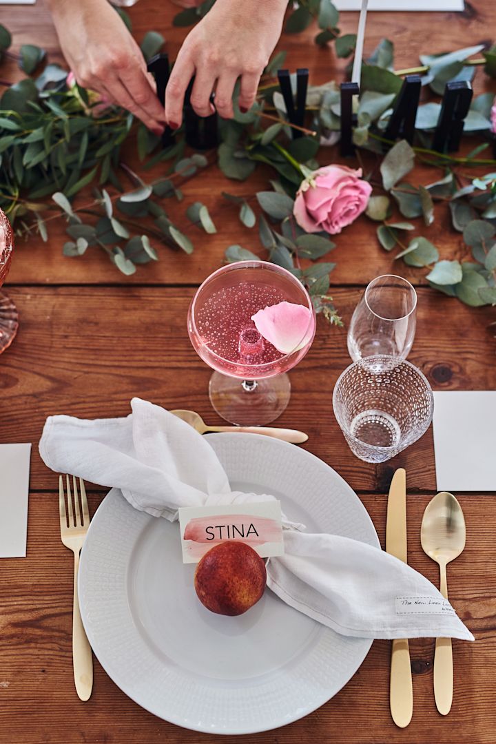 En brunchdukning i rosa toner med eukalyptus som löpare längs med träbordet och Swedish Grace vit tallrik med guldbestick som tallrikskuvert. 
