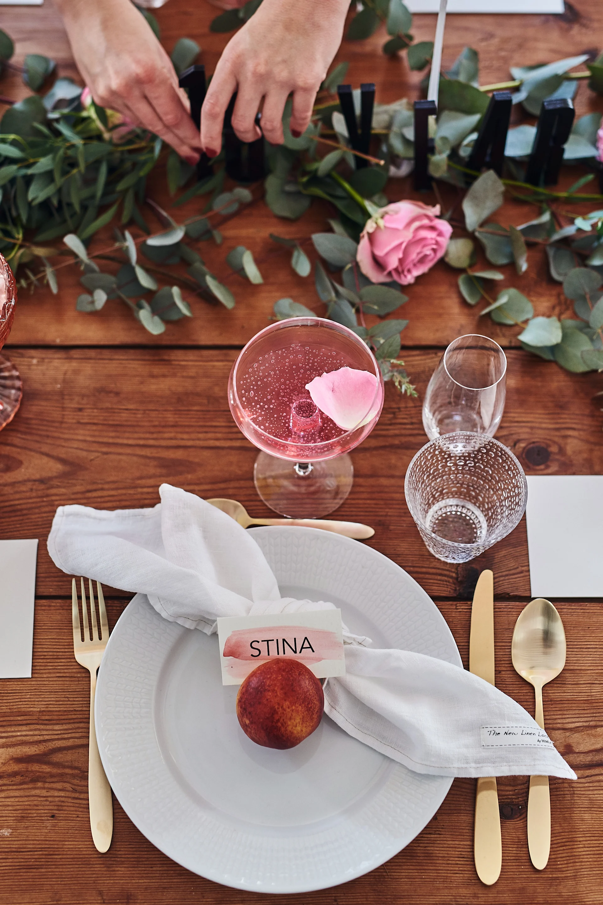 En brunchdukning i rosa toner med eukalyptus som löpare längs med träbordet och Swedish Grace vit tallrik med guldbestick som tallrikskuvert.