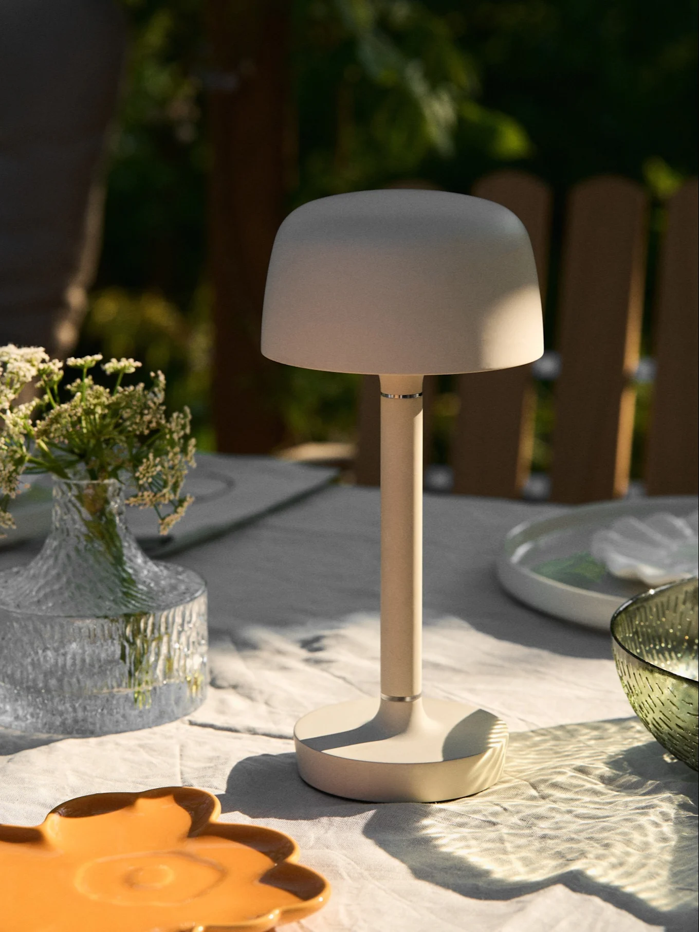 Beige bärbar bordslampa på ett utomhusbord med blomvas och fläckigt solljus.