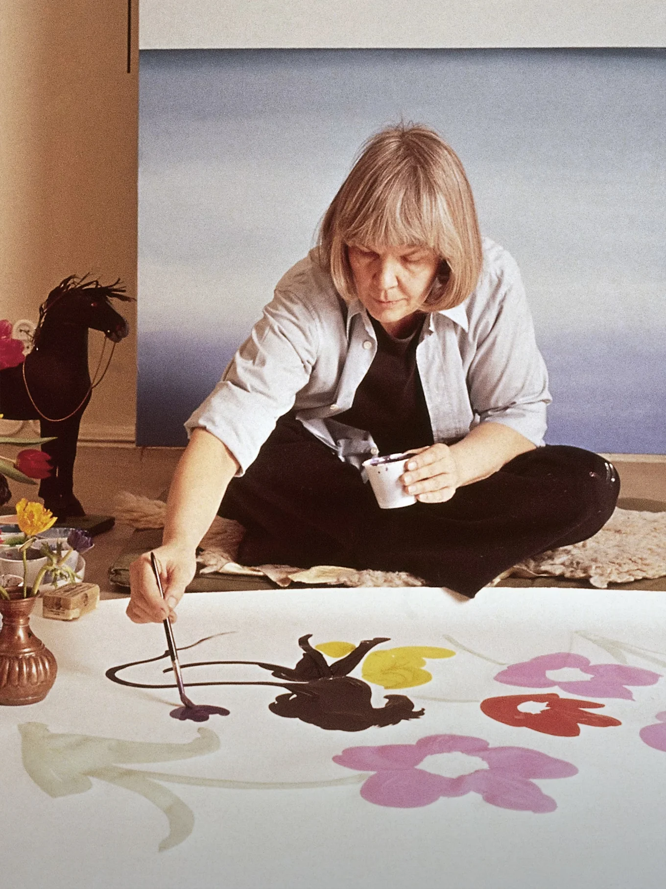 Konstnären Marimekko Maija Isola sitter på golvet och målar stora, färgglada blommor på ett vitt tyg.