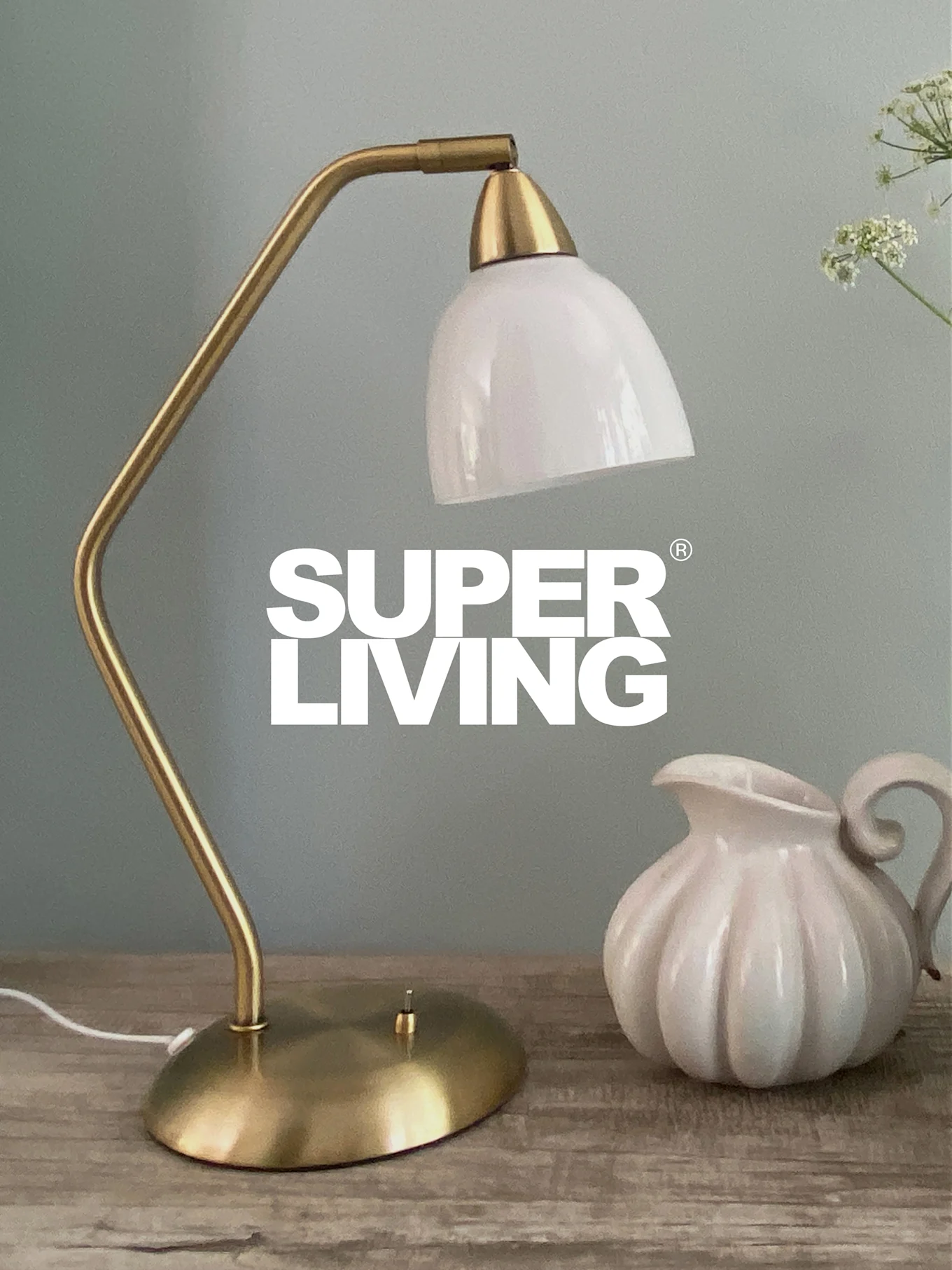 SUPER LIVING-varumärke över en mässingsskrivbordslampa med vit skärm och en vit, räfflad kanna på ett träbord.