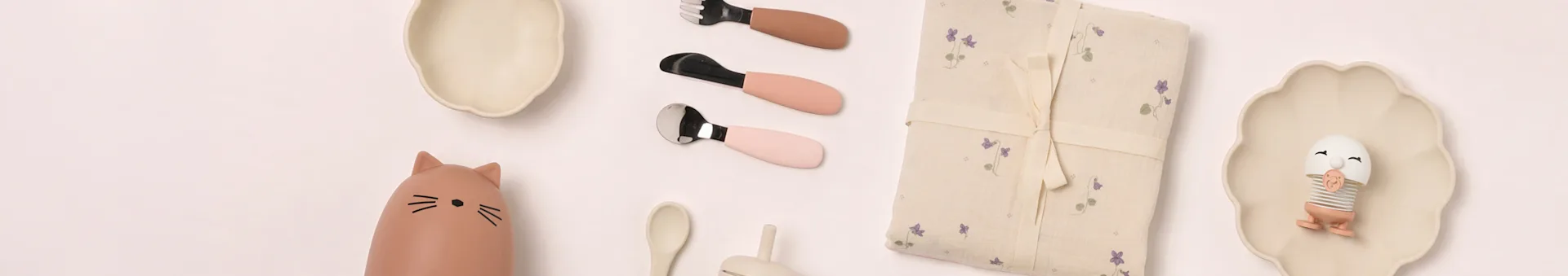 Flat lay av babyprodukter: beige tallrikar, kattflaska, rosa bestick, blommig filt, äggleksak.