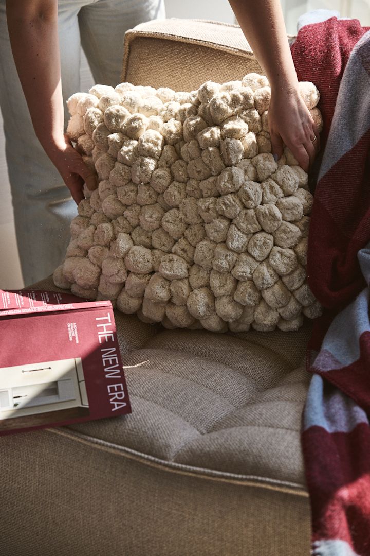 En annan väldigt omtyckt produkt från Byon är Pom pom kudde, här i offwhite i beige soffa med randig pläd i vinrött och ljusblått bredvid.