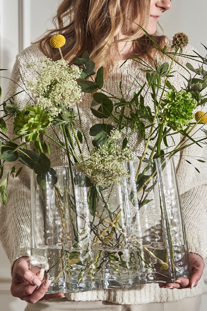 En kvinna i en krämfärgad tröja står och håller i Gry wide vas i färgen clear, som har en bukett vilda vårblommor i sig. 