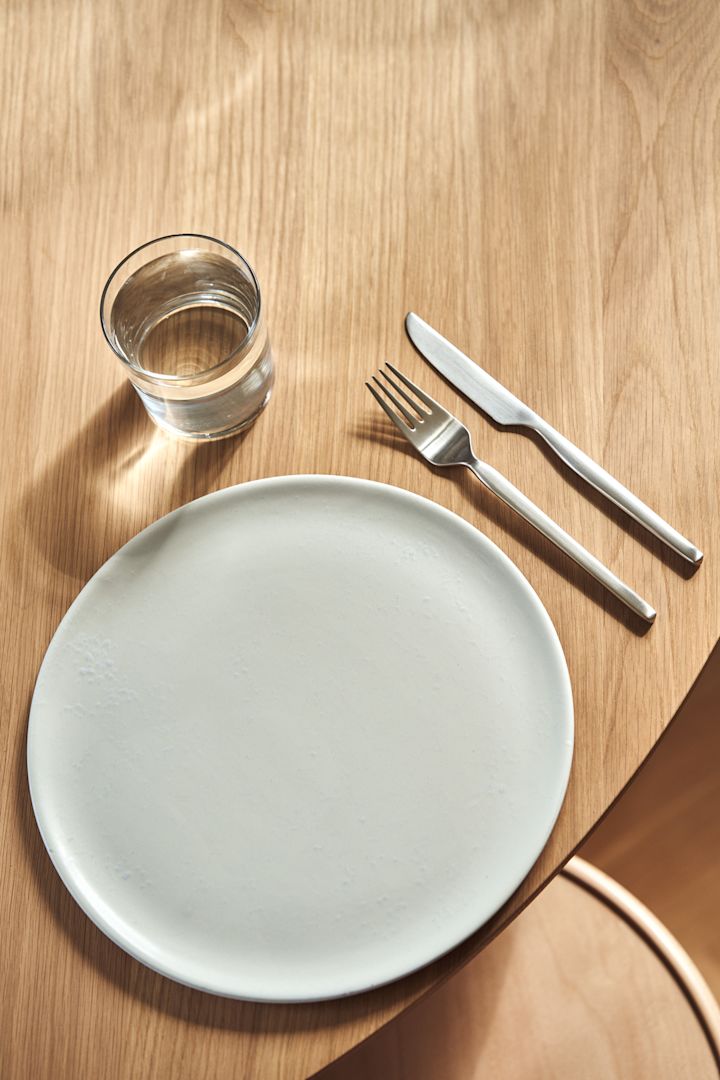 Dorotea-bestick tillsammans med tallrik och glas på träbord, bild tagen uppifrån.